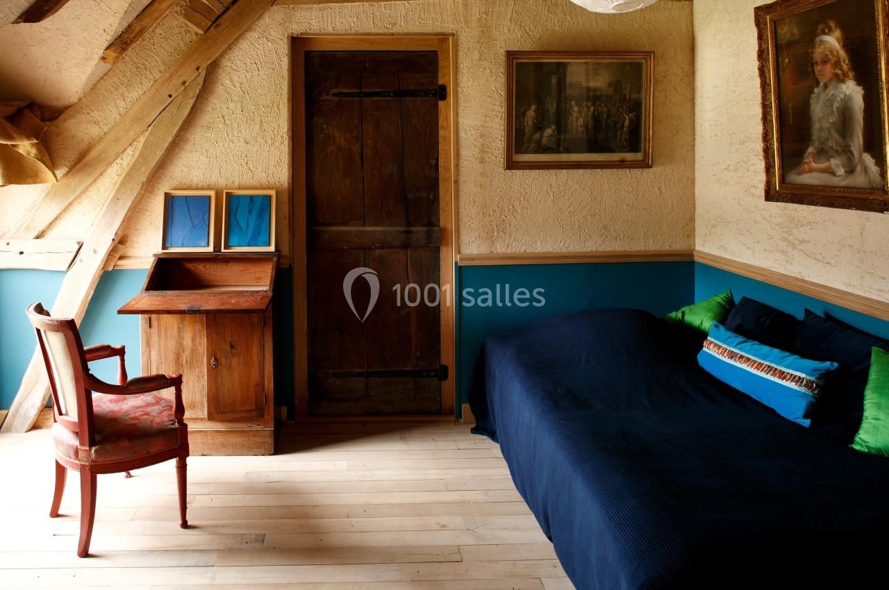 Chambre mansardée avec lit bleu, chaise en bois, bureau ancien, poutres apparentes et tableaux accrochés aux murs.
