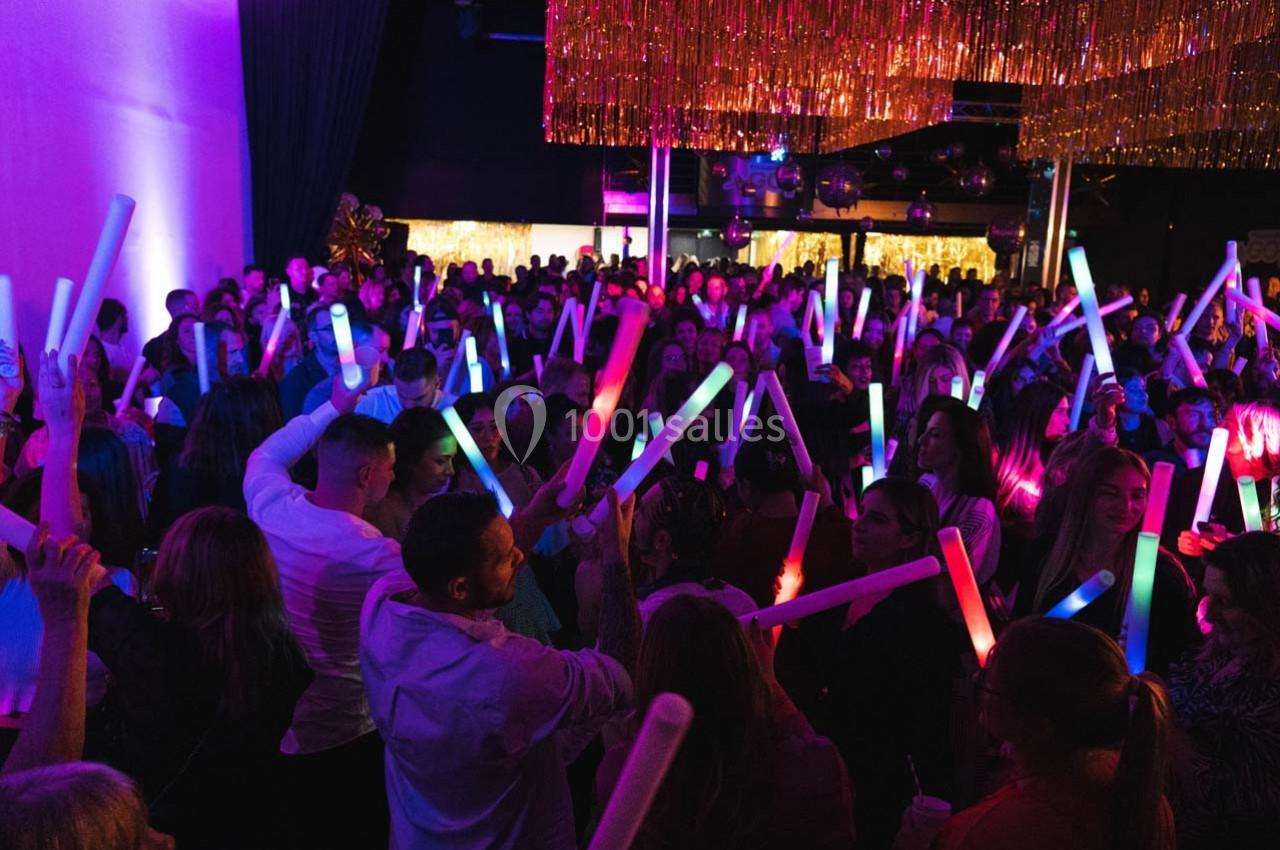 Foule dans une salle sombre tenant des bâtons lumineux colorés sous des décorations dorées.