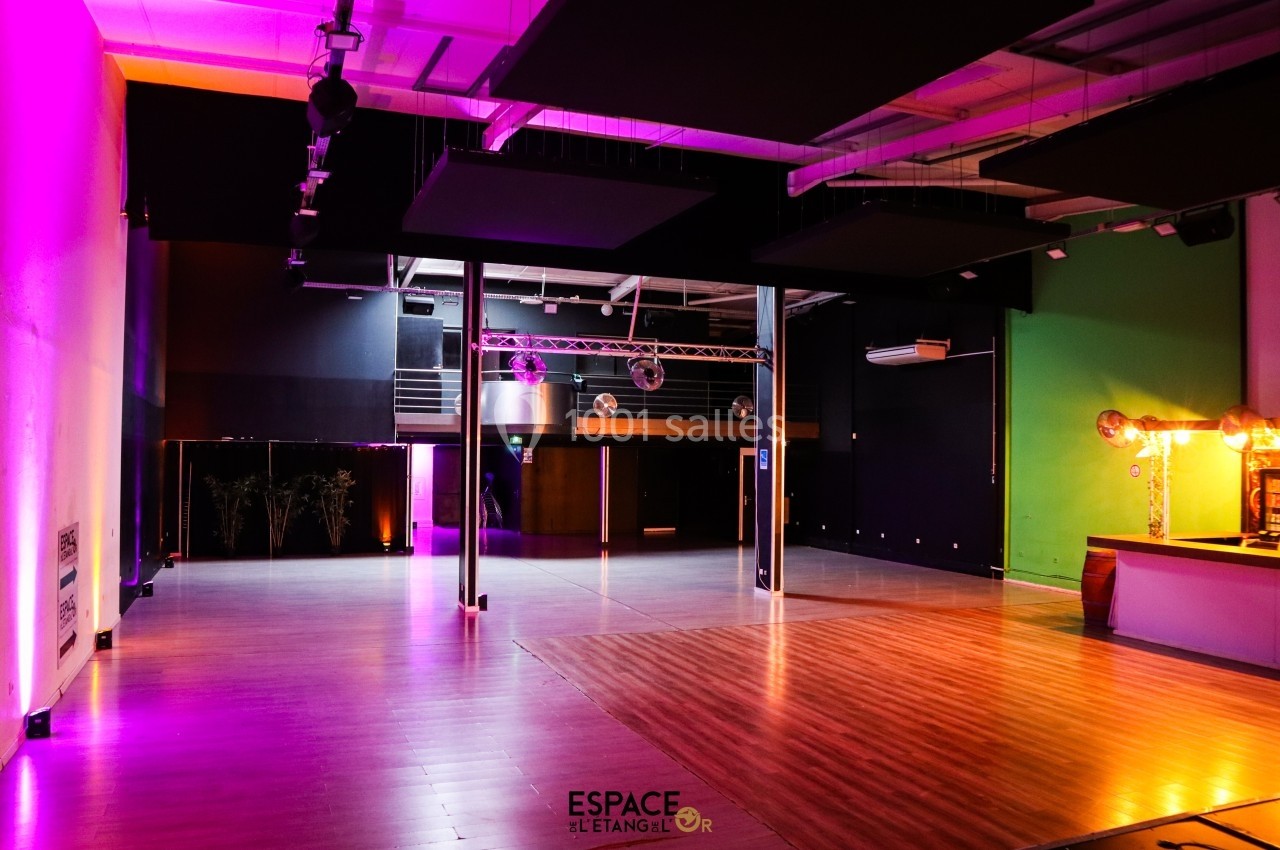 Salle spacieuse avec éclairage coloré, parquet au sol, mezzanine et bar à droite.