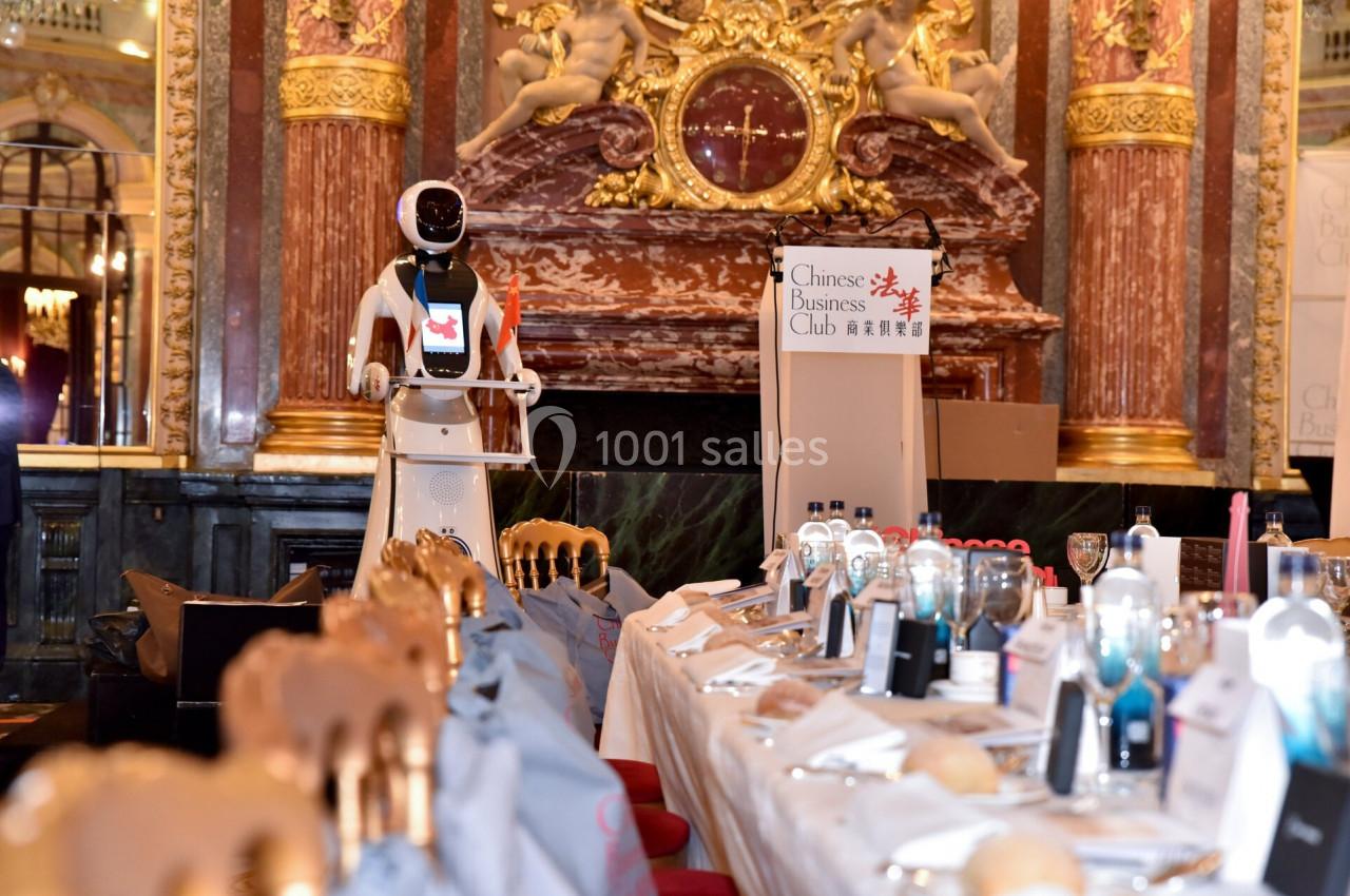 Un robot humanoïde présenté dans une salle ornée, devant une table dressée pour un événement formel.