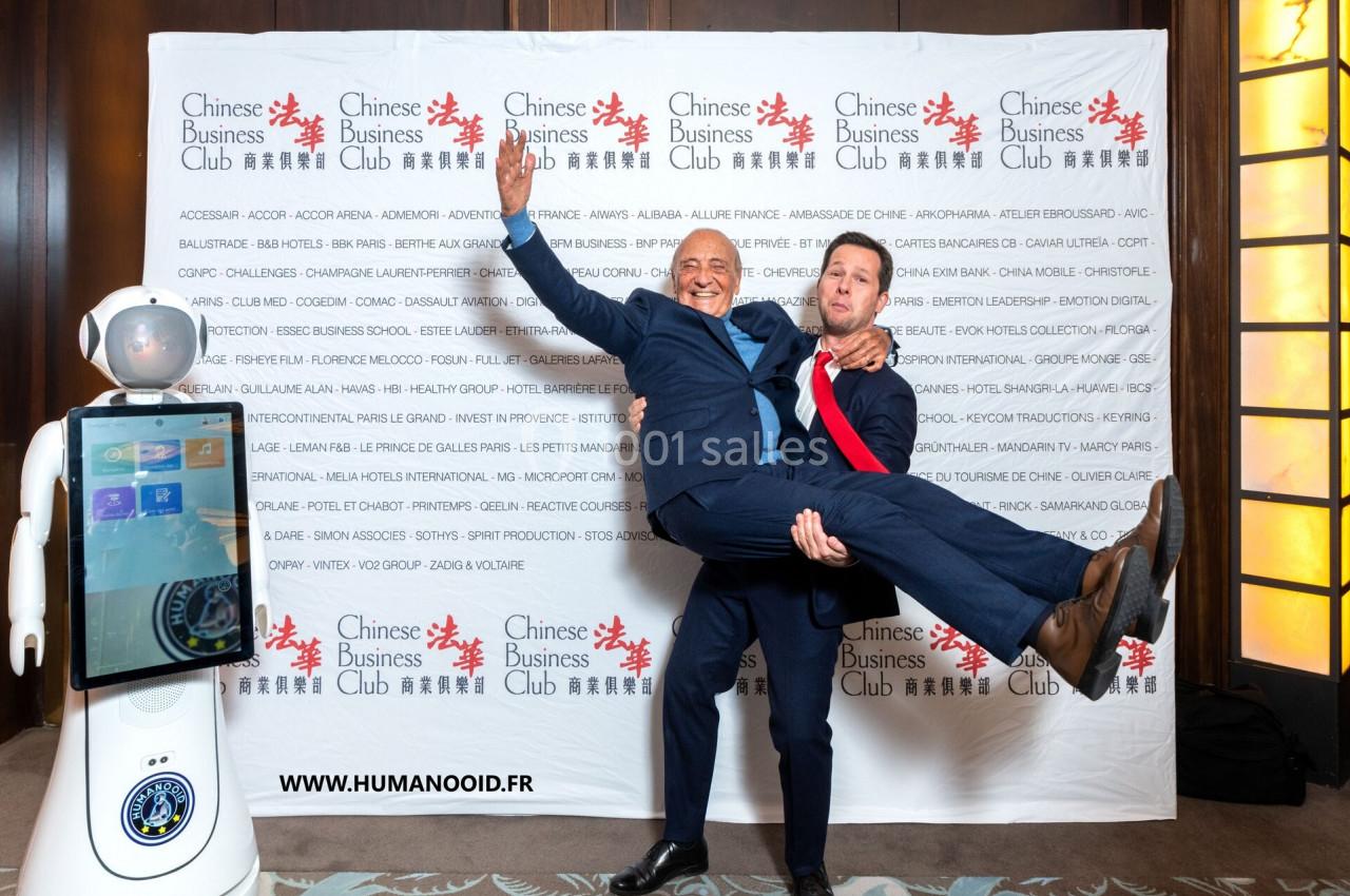 Deux hommes en costume posent de manière joyeuse devant un fond avec des logos, un robot humanoïde à gauche.