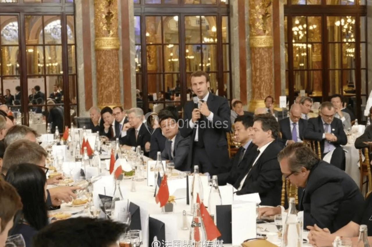 Un homme debout avec un micro s'exprime devant une assemblée lors d'un dîner officiel dans une salle ornée.
