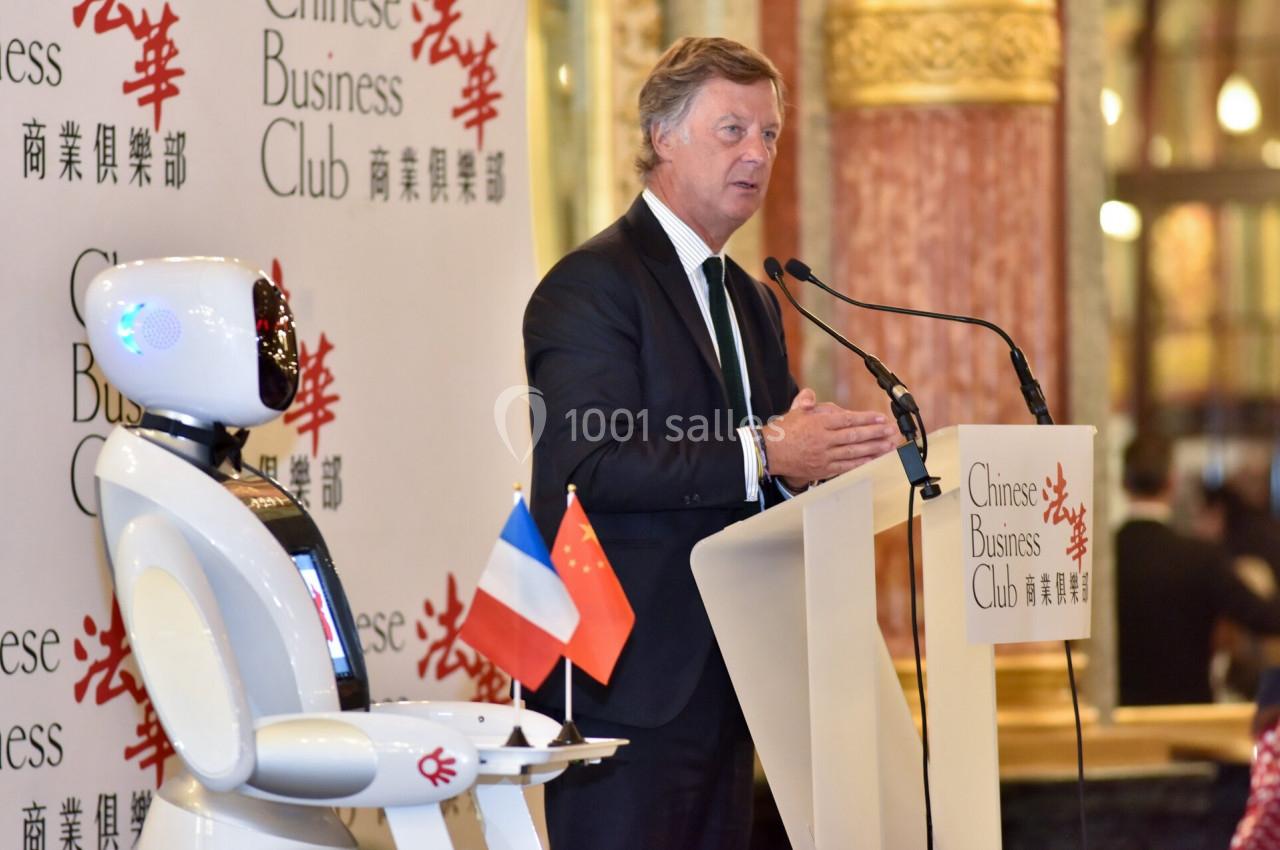 Un homme en costume parle à un pupitre avec un robot à ses côtés, dans un cadre formel décoré de drapeaux français et…