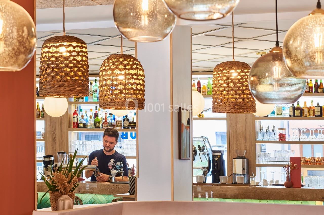 Un barman prépare une boisson derrière un comptoir, entouré de bouteilles et de suspensions lumineuses décoratives.