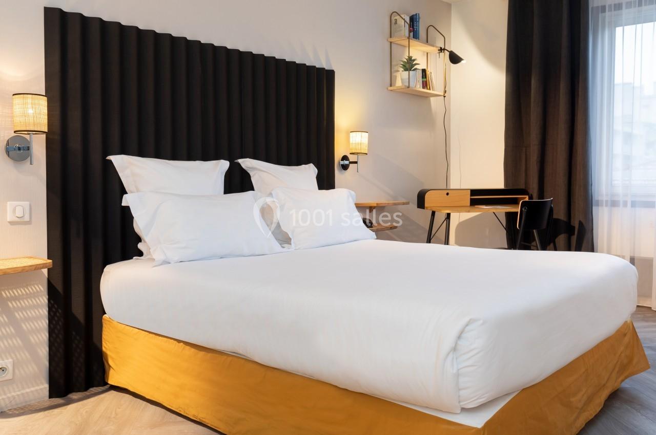 Lit double avec draps blancs, tête de lit noire, bureau en bois avec chaise et étagères murales dans une chambre lumineuse.