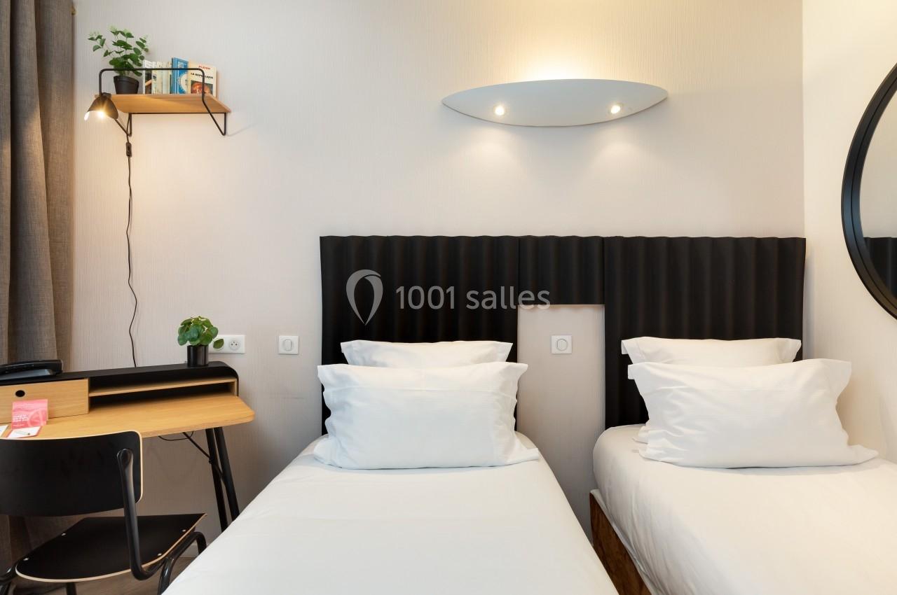 Deux lits simples avec tête de lit noire, bureau en bois, chaise noire et étagère murale avec plante.