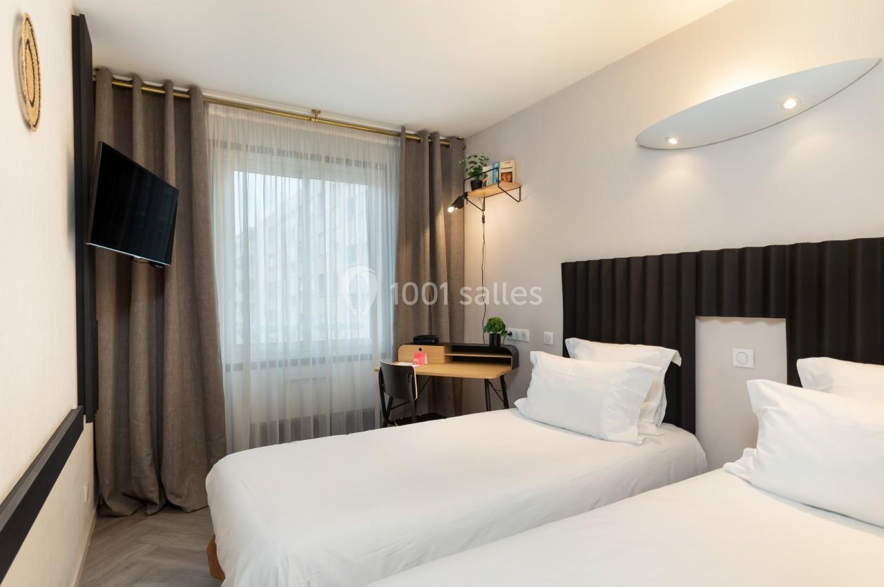 Chambre lumineuse avec deux lits simples, bureau, télévision murale et grande fenêtre avec rideaux.