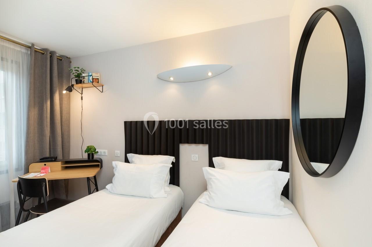 Deux lits simples avec tête de lit noire, bureau en bois, chaise noire, miroir rond et lumière naturelle.