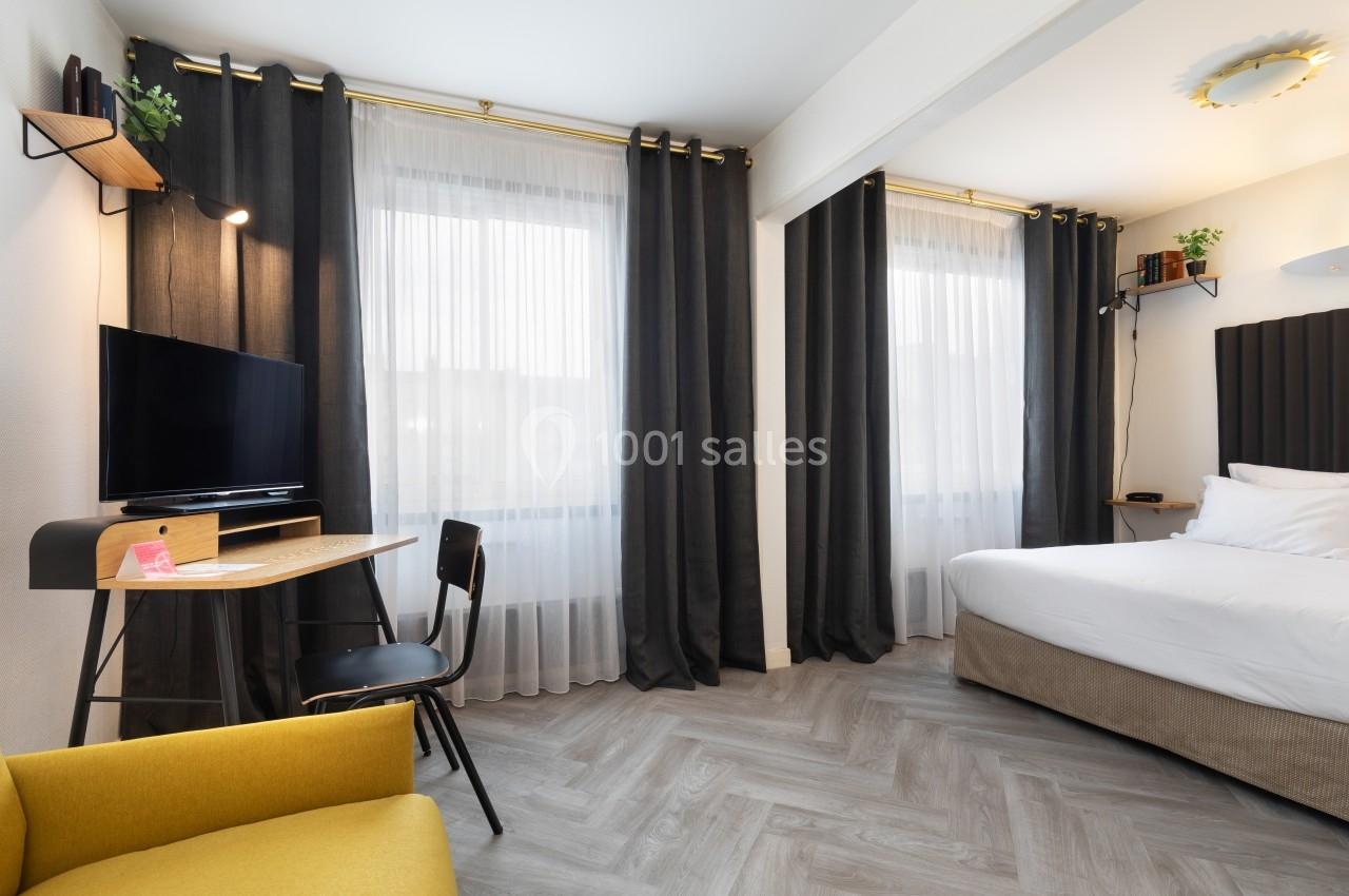 Chambre lumineuse avec lit double, bureau équipé, télévision, fauteuil jaune et rideaux sombres devant des fenêtres.