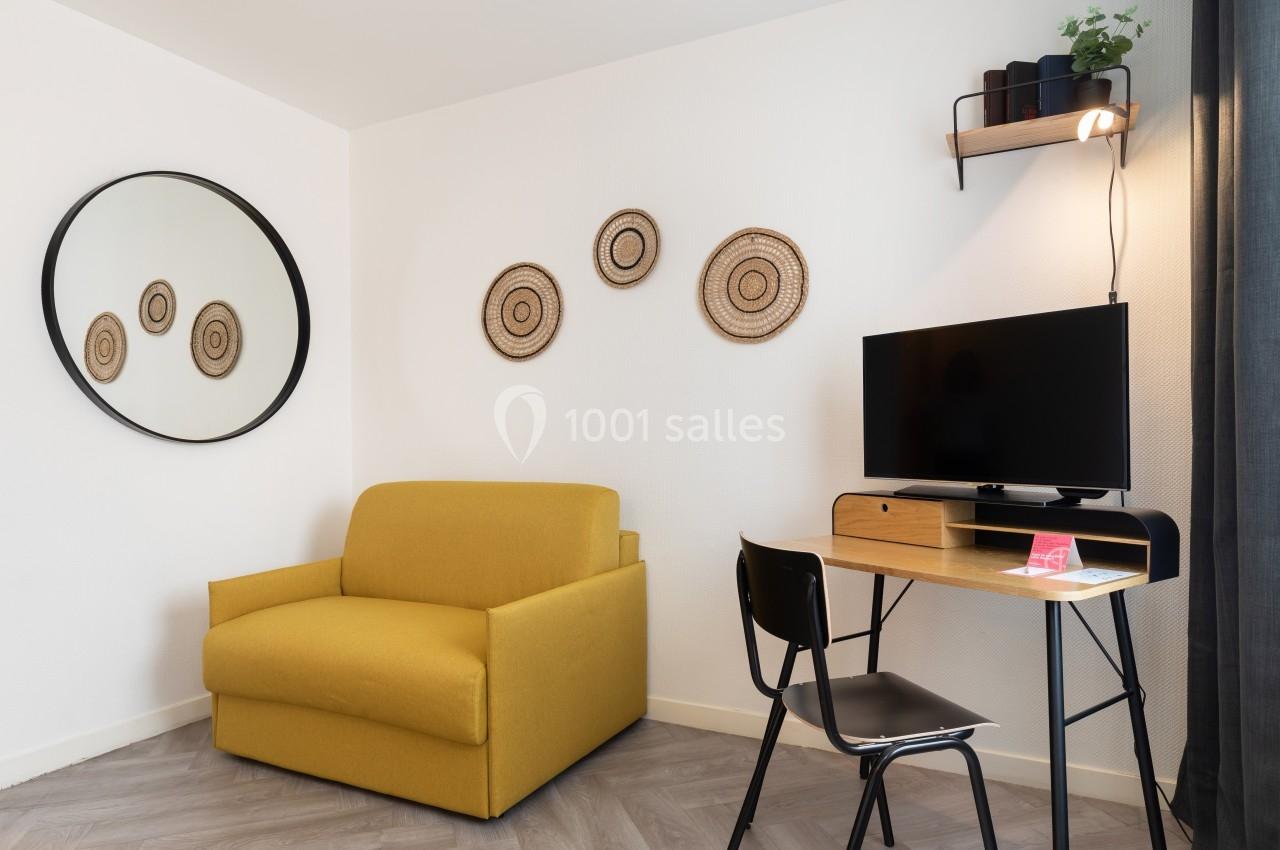 Fauteuil jaune, bureau en bois avec chaise noire, télévision, miroir rond et décorations murales en osier.