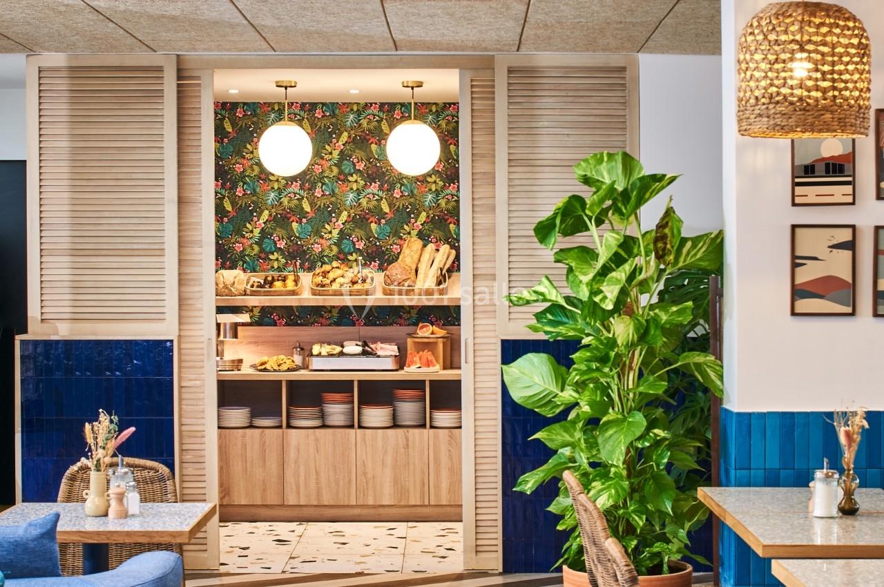 Espace intérieur lumineux avec mobilier en rotin, plantes vertes et vue sur un buffet décoré d'un papier peint floral.