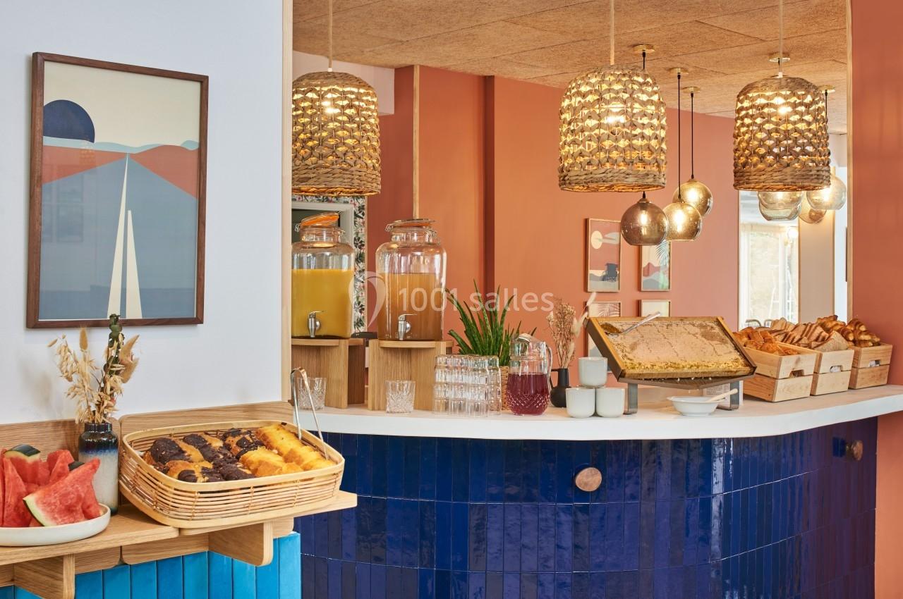 Buffet de petit-déjeuner avec viennoiseries, fruits, boissons et décor chaleureux aux tons bleus et orangés.