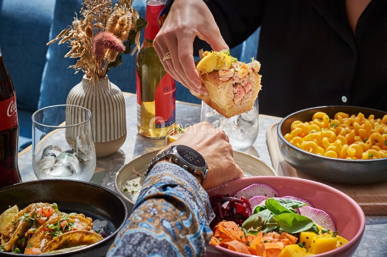 Repas convivial avec plats variés, dont pâtes, salade colorée et sandwich, sur une table décorée de fleurs séchées.