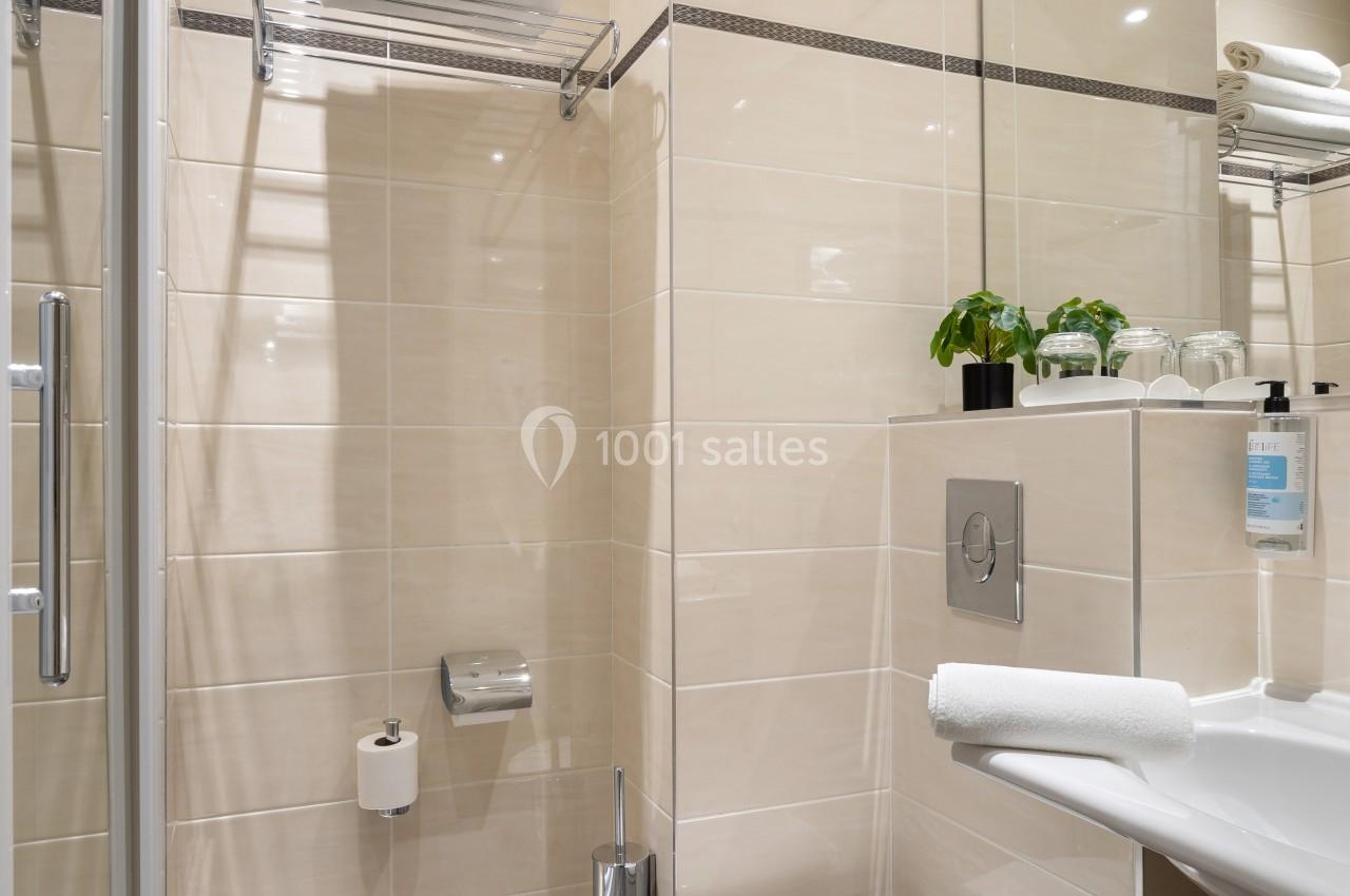 Salle de bain moderne avec douche vitrée, toilettes suspendues, lavabo, serviettes pliées et plantes décoratives.