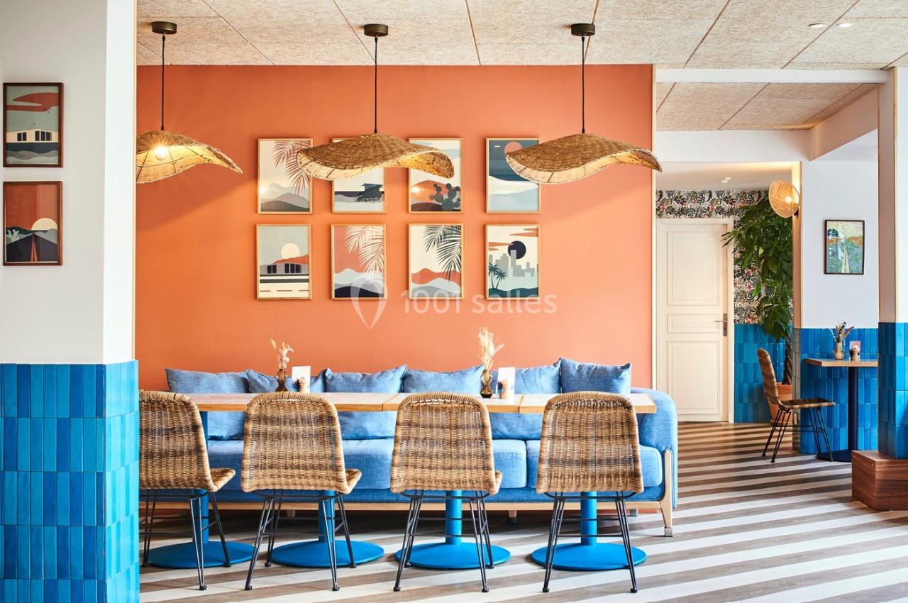 Salle à manger lumineuse avec murs orange, chaises en rotin, table en bois et décorations murales colorées.