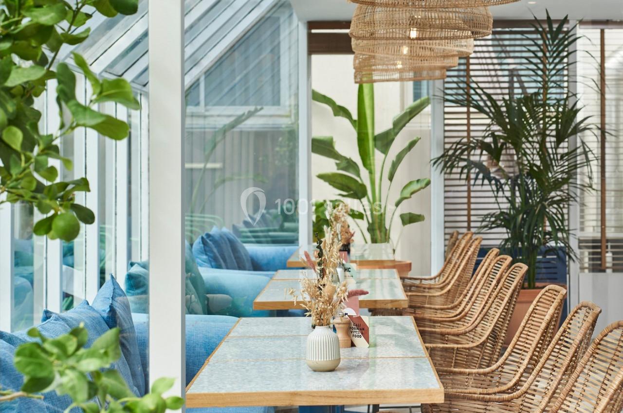 Salle lumineuse avec tables en bois, chaises en rotin, plantes vertes et grandes baies vitrées.