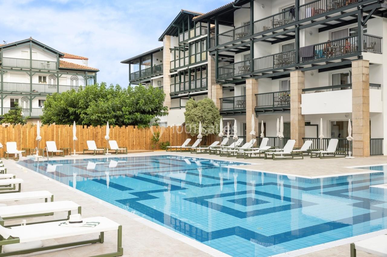Piscine extérieure entourée de chaises longues et d'un bâtiment à balcons en bois, sous un ciel dégagé.