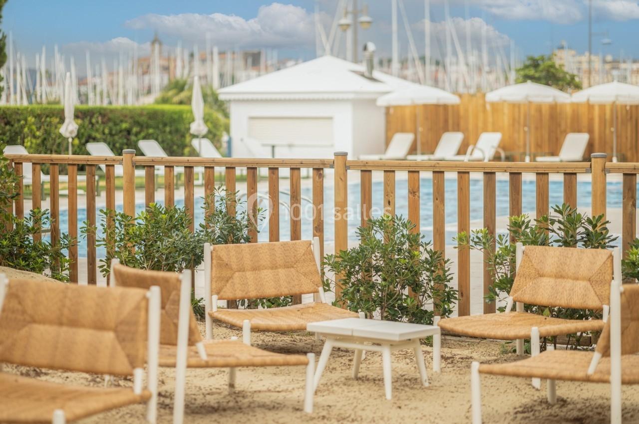 Chaises en rotin disposées autour d'une table basse près d'une piscine, avec une clôture en bois et des voiliers en arrière…
