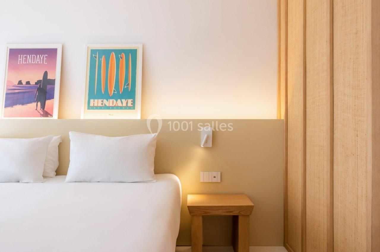 Lit double avec draps blancs, table de chevet en bois et affiches encadrées sur un mur clair dans une chambre lumineuse.