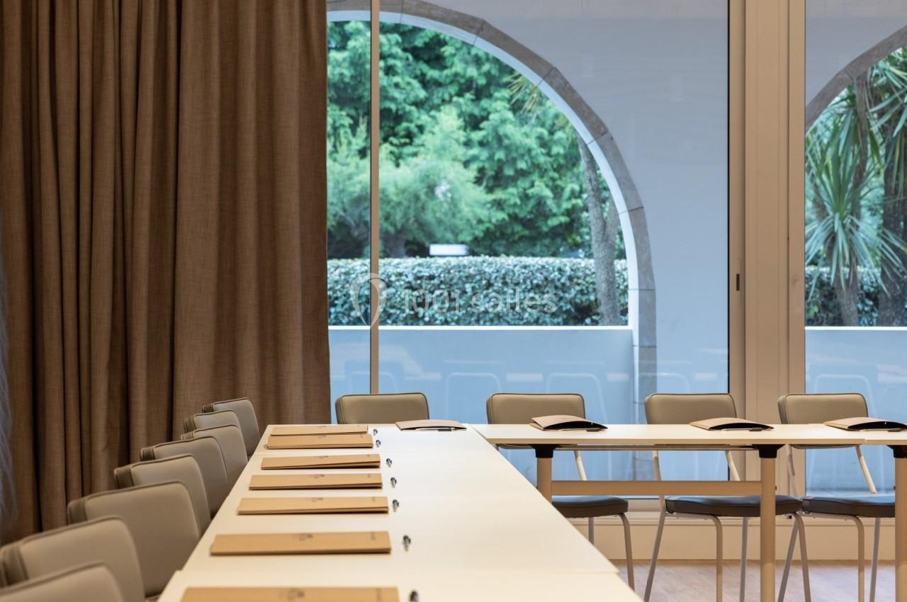 Salle de réunion lumineuse avec des tables blanches alignées, des chaises grises et une grande baie vitrée donnant sur un…