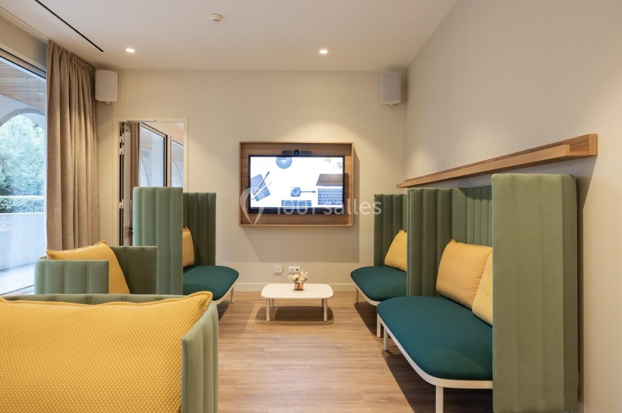 Salle moderne avec canapés verts et jaunes, table basse centrale, écran mural et grande baie vitrée à gauche.