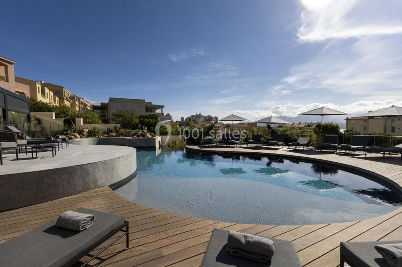 Piscine extérieure avec terrasse en bois, transats et parasols, entourée de bâtiments sous un ciel ensoleillé.