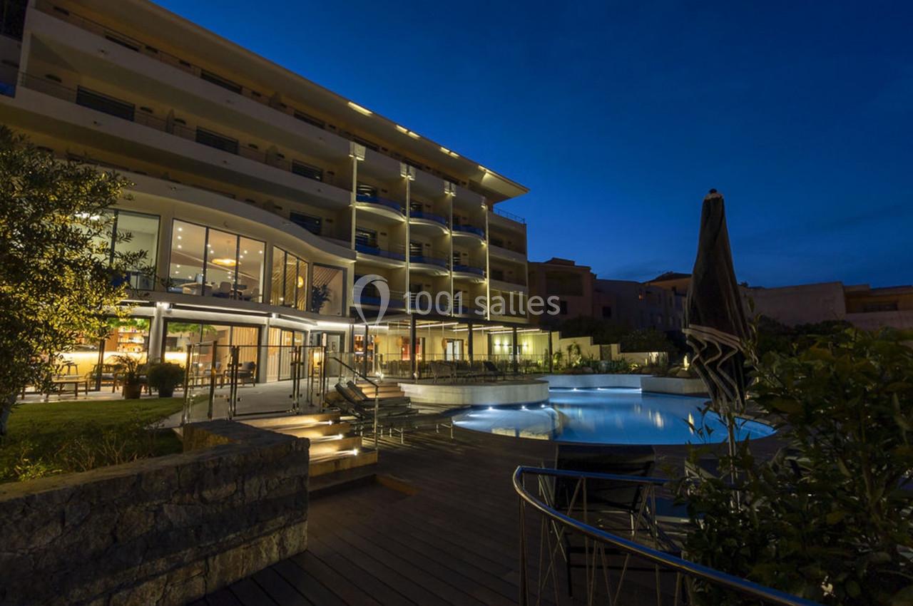 Hôtel moderne éclairé de nuit avec piscine extérieure et terrasse en bois entourée de végétation.