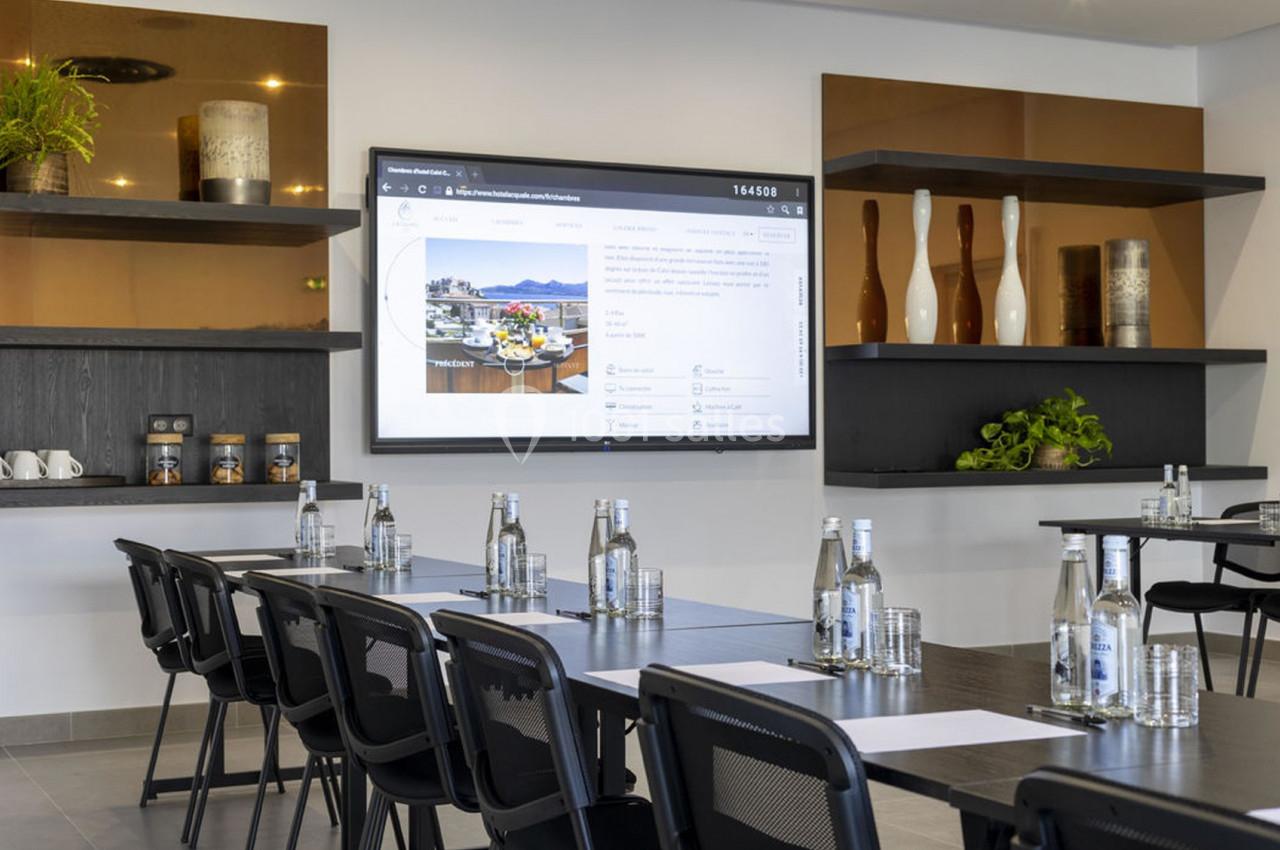 Salle de réunion moderne avec tables noires, chaises, bouteilles d'eau, écran mural et étagères décoratives.