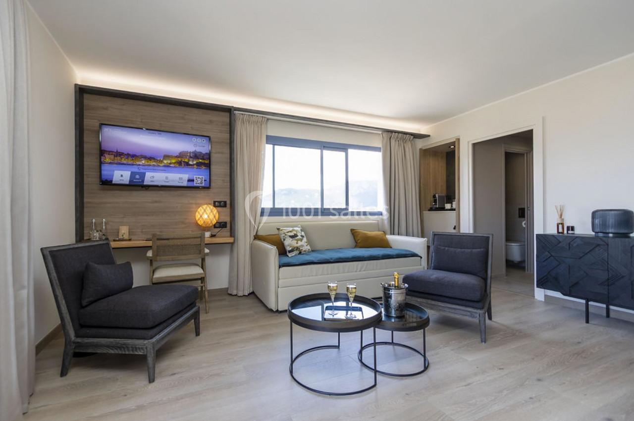 Salon moderne avec canapé-lit, fauteuils, table basse, télévision murale et grande fenêtre laissant entrer la lumière…