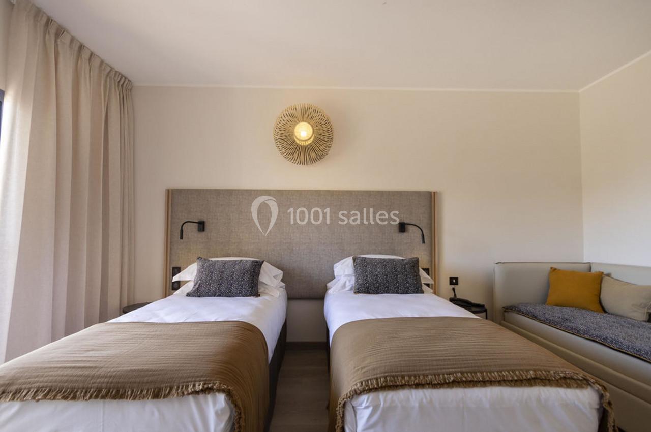 Deux lits simples avec des couvre-lits beige, une applique murale et un canapé avec coussins dans une chambre lumineuse.