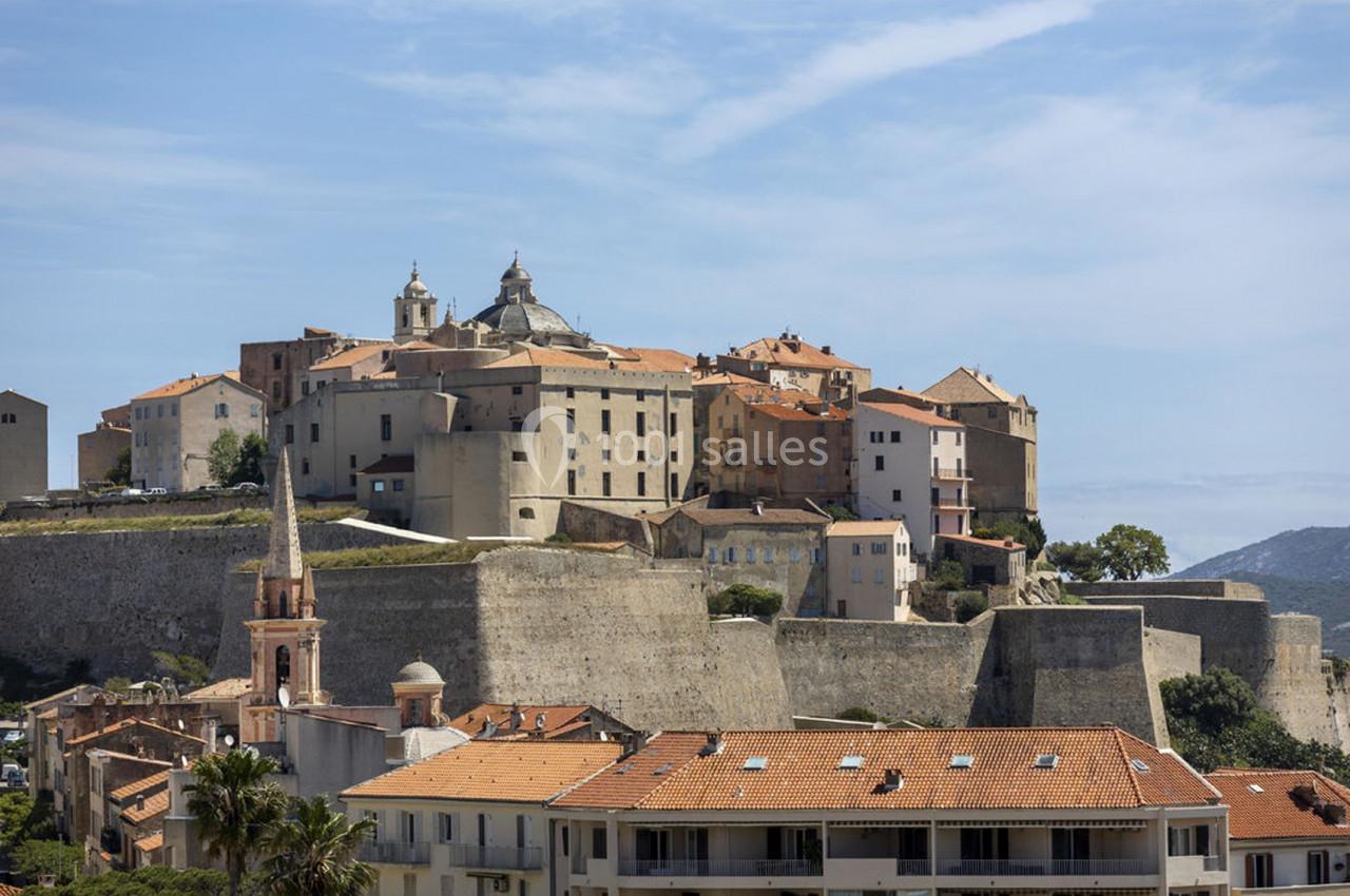 Vue d'une citadelle en pierre sur une colline, entourée de bâtiments aux toits en tuiles et d'un ciel dégagé.