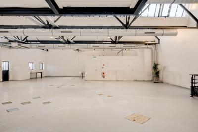 Intérieur d'un grand espace industriel lumineux avec sol clair, murs blancs, poutres métalliques et conduits apparents.