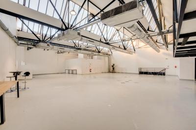 Intérieur d'un grand espace industriel lumineux avec sol clair, murs blancs, poutres métalliques et conduits apparents.