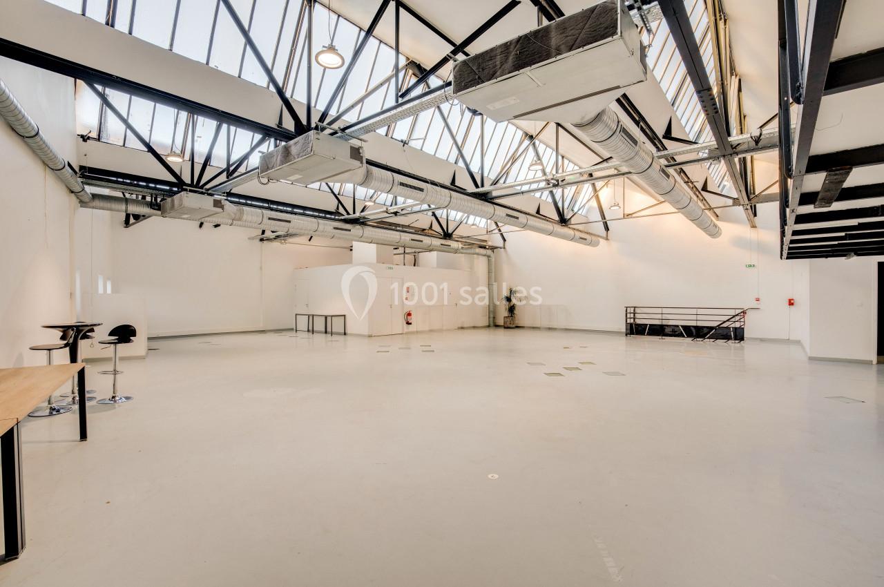 Grand espace intérieur lumineux avec plafond en verrière, poutres métalliques apparentes et sol en béton clair.