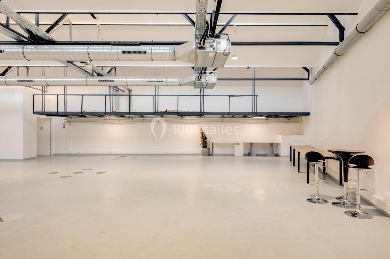 Intérieur d'un espace industriel lumineux avec plafond haut, conduits apparents et mobilier minimaliste.