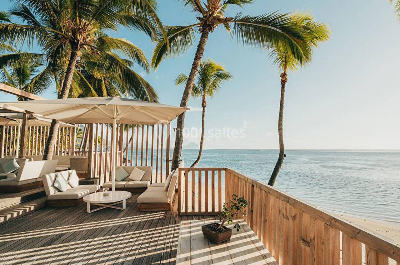Terrasse en bois avec fauteuils et parasol, surplombant une plage bordée de palmiers et l'océan sous un ciel dégagé.
