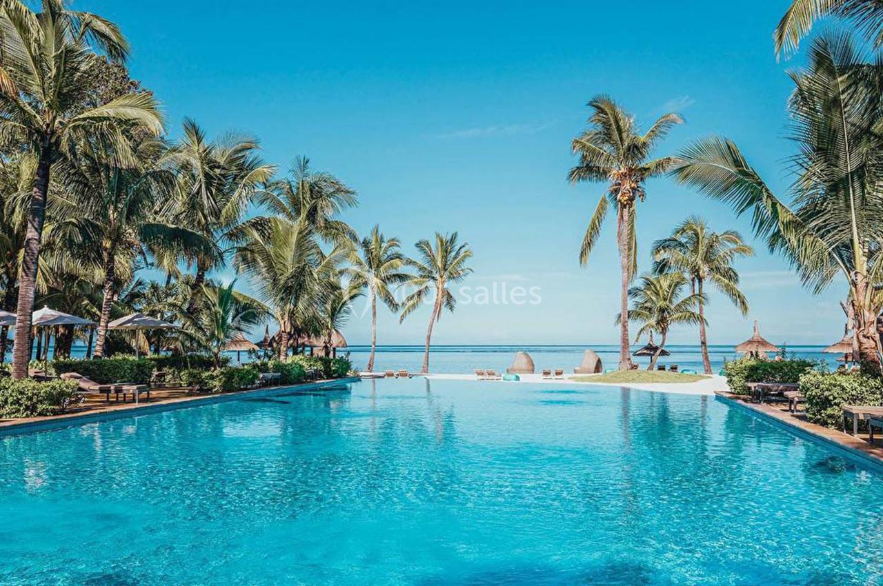 Piscine extérieure entourée de palmiers avec vue sur la mer sous un ciel bleu clair.