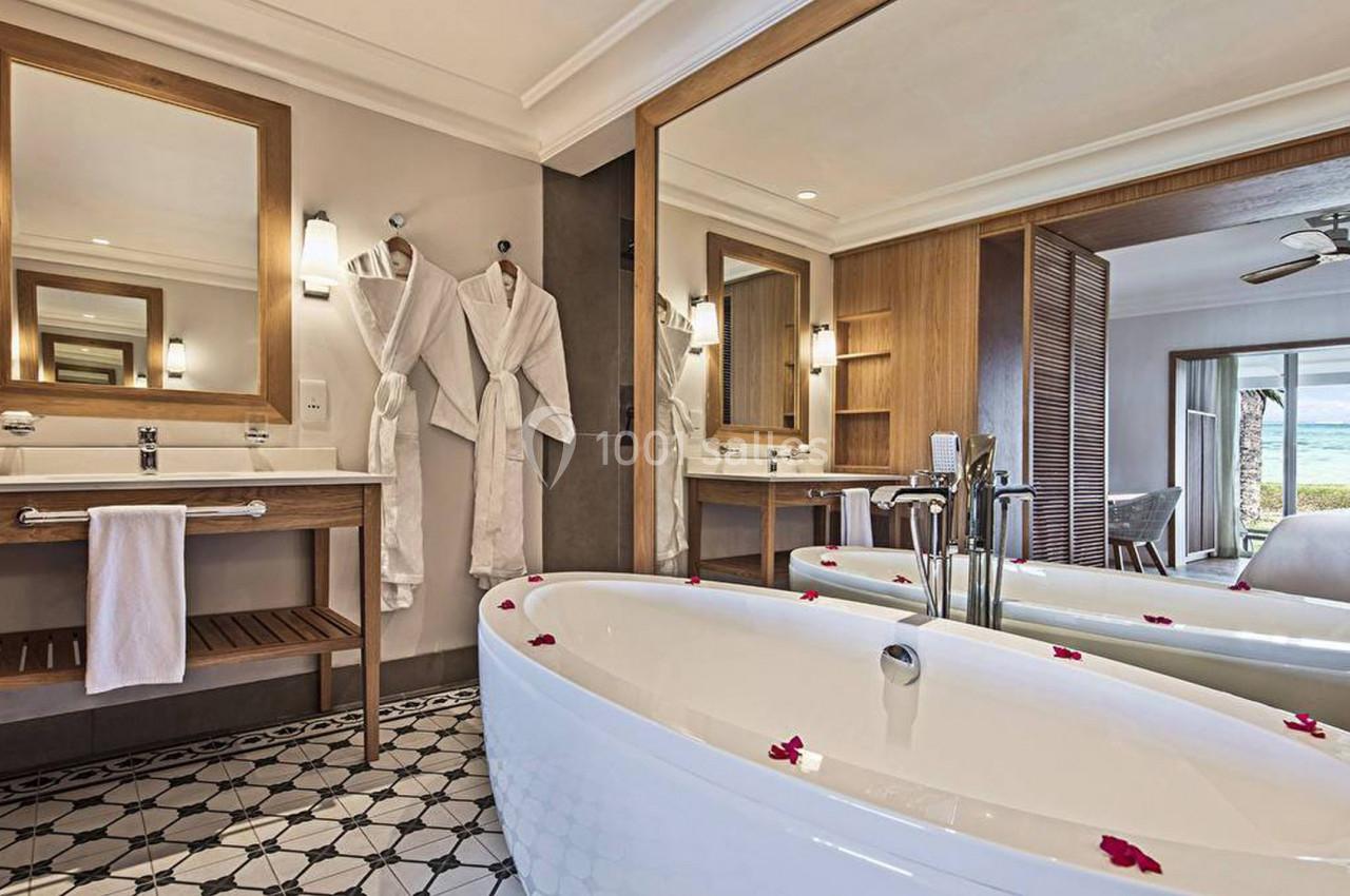 Salle de bain lumineuse avec baignoire blanche, miroirs, peignoirs accrochés et vue sur l'extérieur.