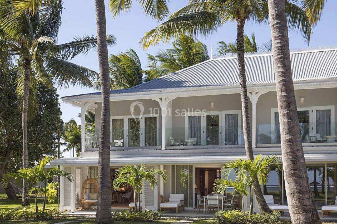 Maison à deux étages entourée de palmiers, avec une façade blanche, un toit en tôle et une terrasse meublée.