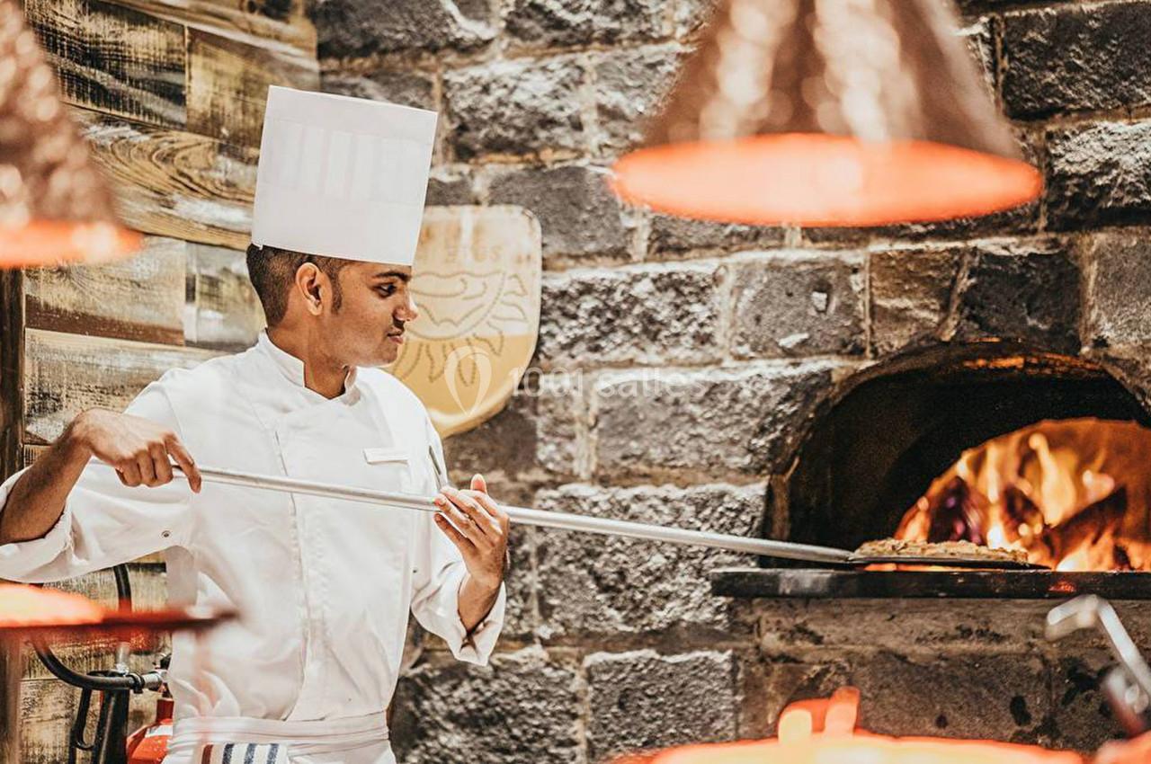 Un chef en tenue blanche enfourne une pizza dans un four à bois en pierre dans une cuisine rustique.