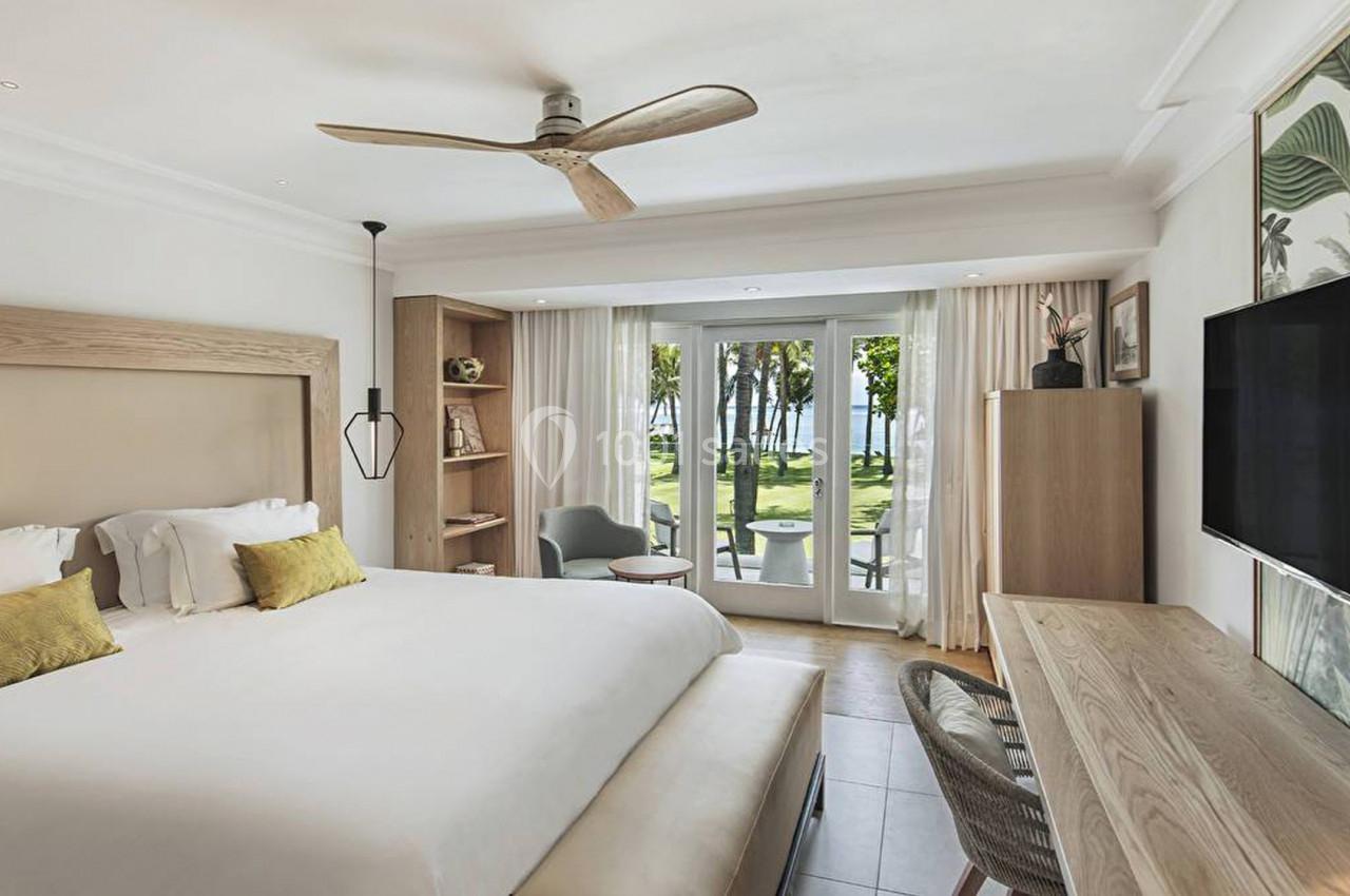 Chambre lumineuse avec lit double, bureau, télévision, ventilateur au plafond et vue sur un jardin menant à la mer.