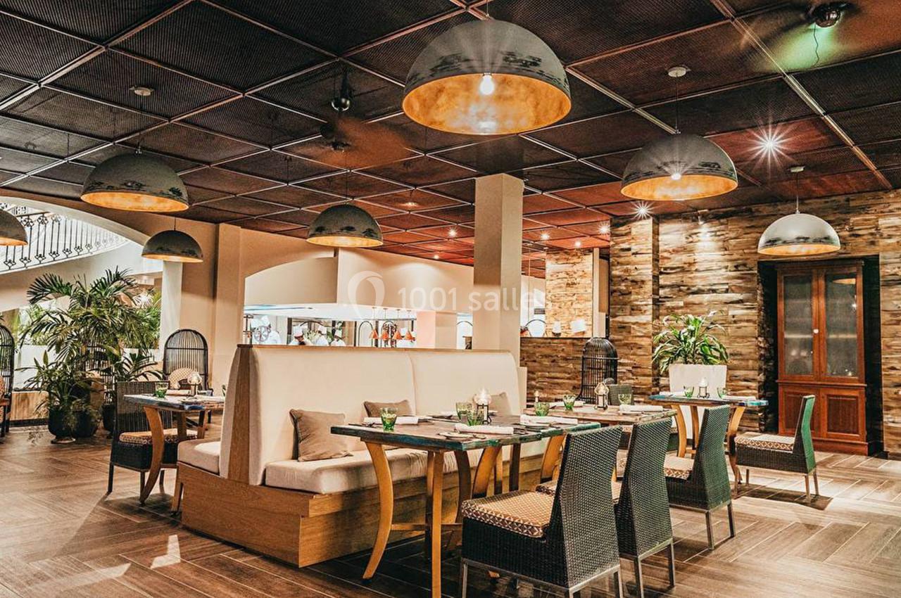 Salle de restaurant élégante avec tables dressées, chaises en osier, éclairage suspendu et décoration boisée.