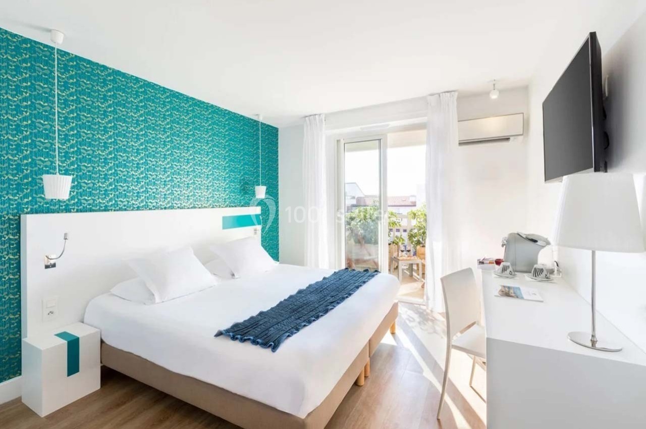 Chambre lumineuse avec lit double, bureau, télévision murale, balcon et décoration murale turquoise.