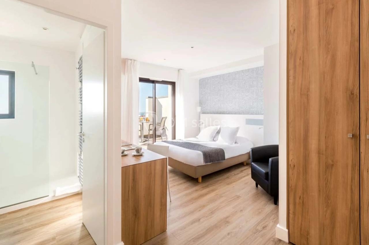 Chambre lumineuse avec lit double, sol en bois, salle de bain attenante et balcon donnant sur l'extérieur.