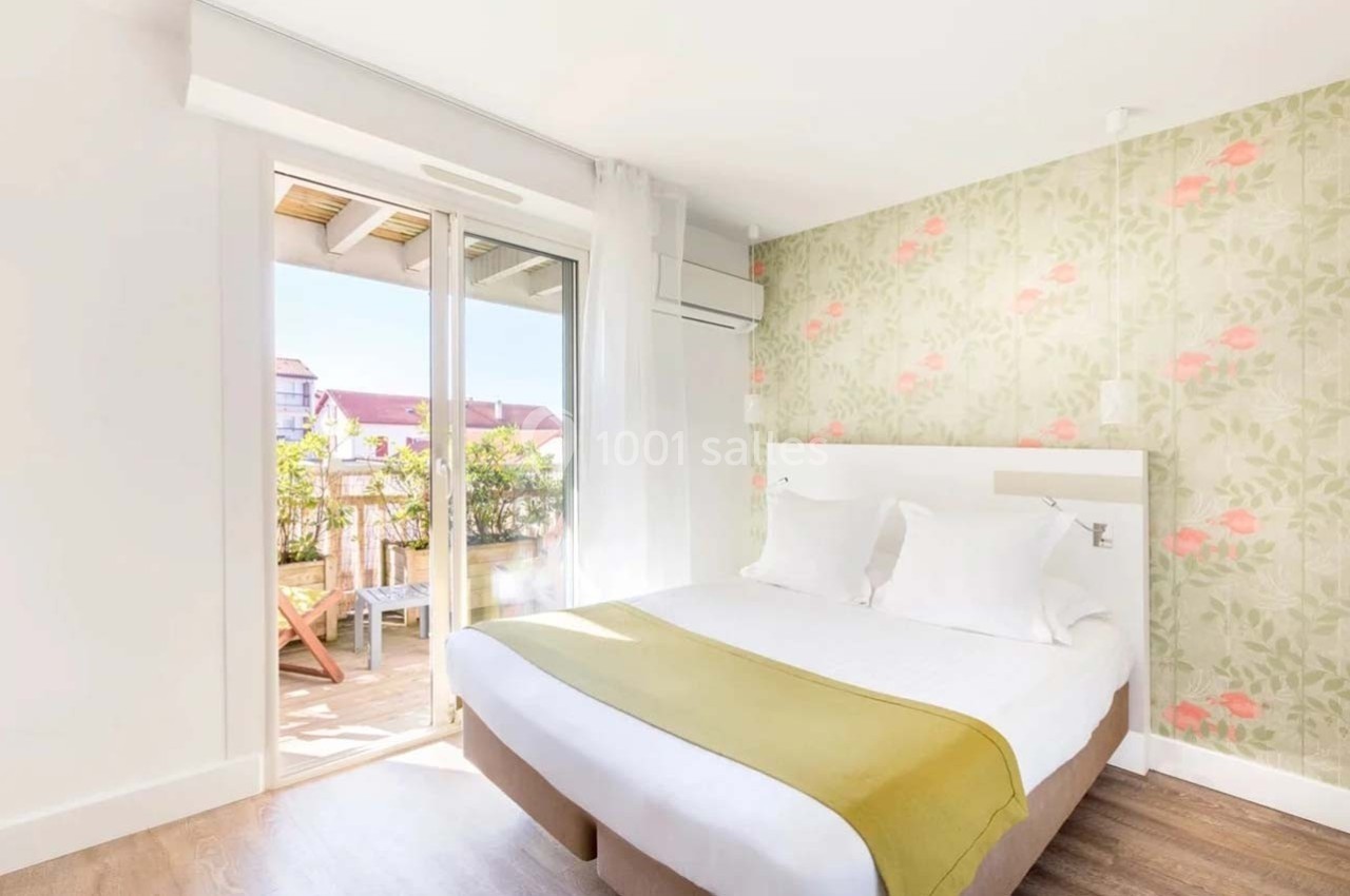 Chambre lumineuse avec lit double, mur à motif floral, parquet et accès à un balcon aménagé.