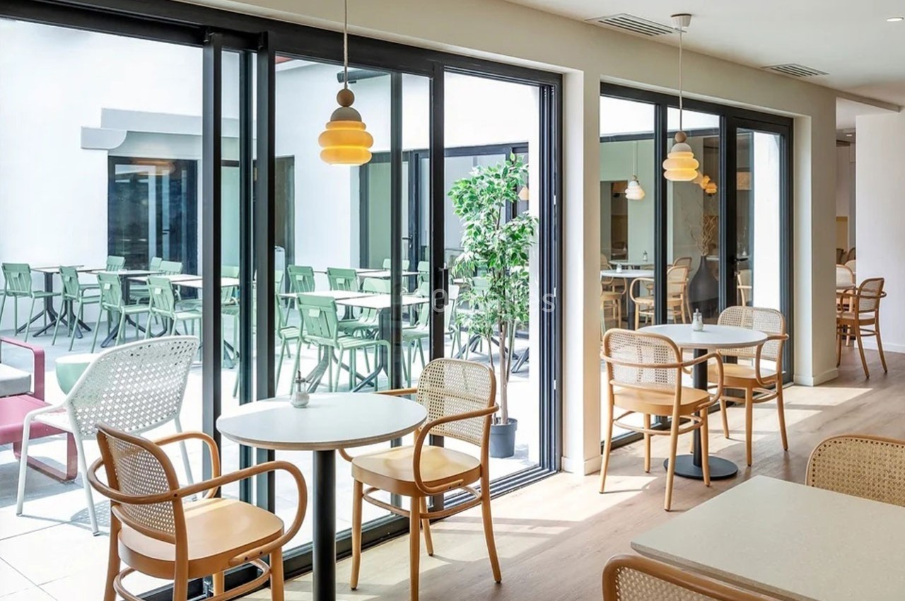 Salle lumineuse avec tables et chaises en bois, grandes baies vitrées donnant sur une terrasse avec mobilier extérieur.