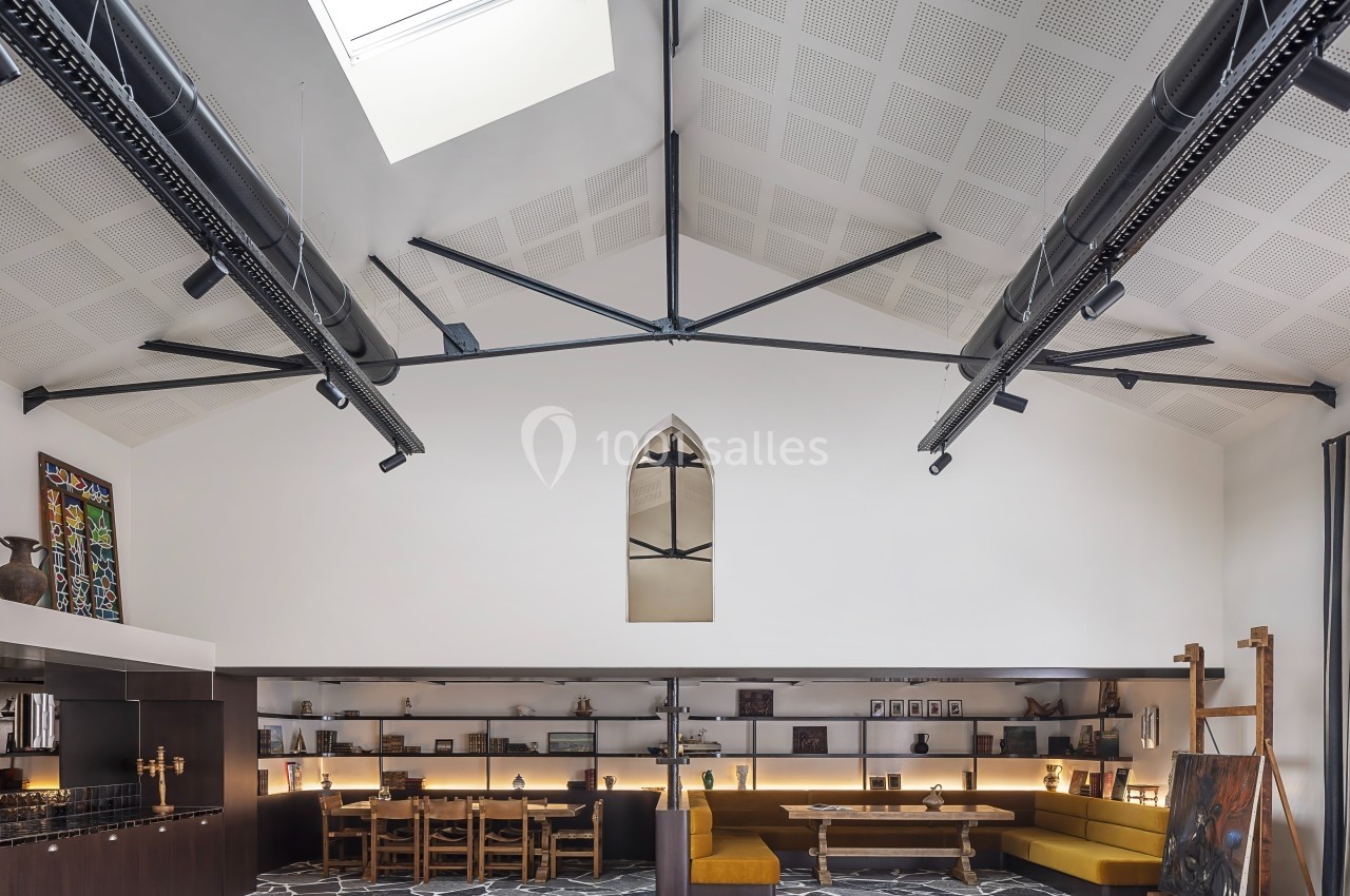 Salle lumineuse avec toit voûté, poutres apparentes, tables en bois, banquettes jaunes et sol en pierre.