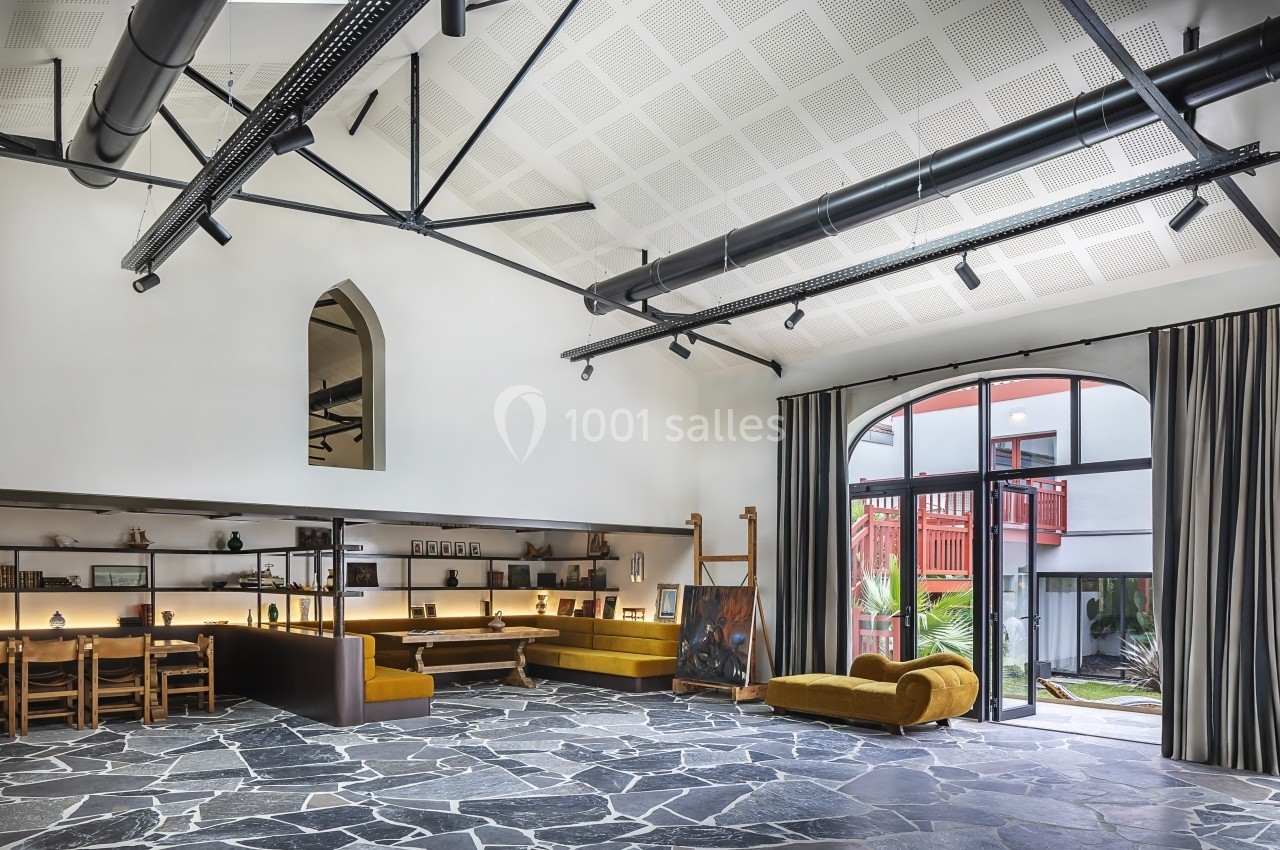 Salle lumineuse avec plafond haut, poutres apparentes, mobilier en bois, canapés et grande baie vitrée donnant sur un patio.