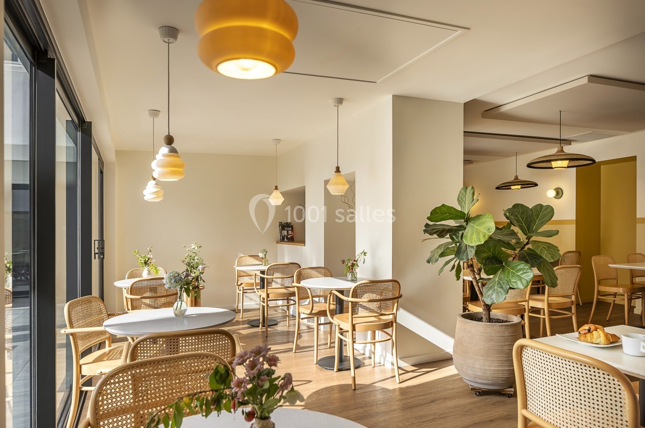 Salle lumineuse avec tables rondes, chaises en rotin, plantes décoratives et grandes fenêtres laissant entrer la lumière…