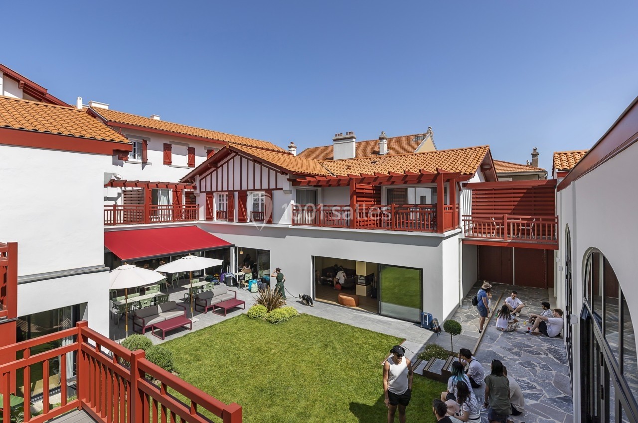 Cour intérieure d'un bâtiment aux façades blanches et rouges, avec terrasse, parasols et personnes assises.